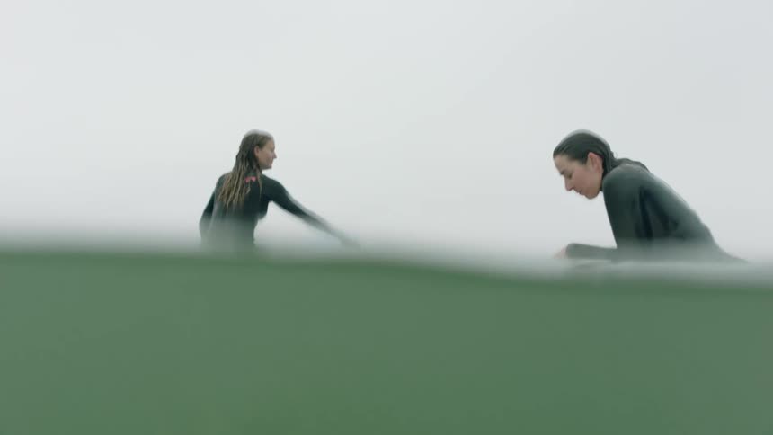 A serene, slow-motion, water-level shot shows two female surfers in wetsuits patiently waiting for waves on a foggy day