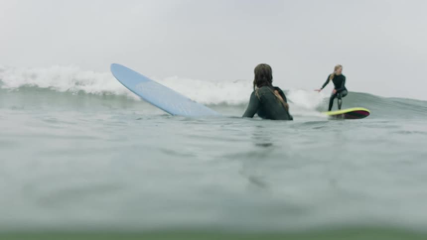 A woman waits on her surfboard in the ocean while another surfer rides a wave in the background on an overcast day