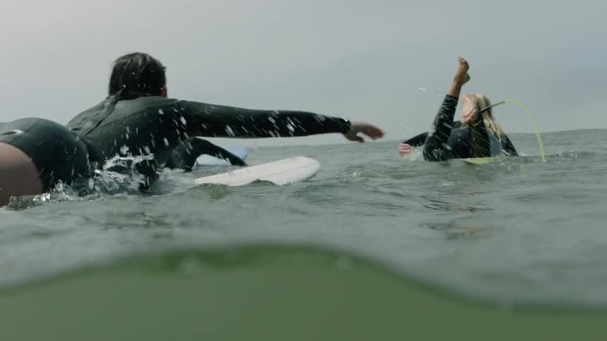 A slow-motion, immersive water-level view of a group of friends paddling out on their surfboards on a calm, overcast day