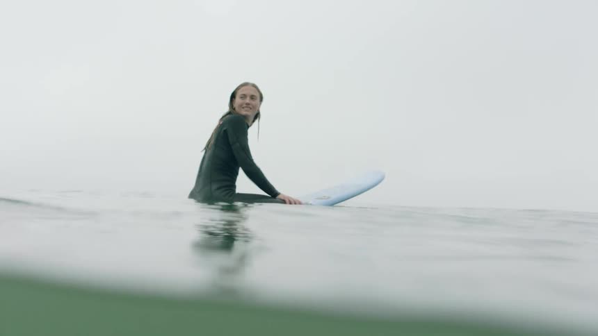 A smiling female surfer in a black wetsuit waits on her board in the calm, foggy ocean, shot in slow motion from the water's surface