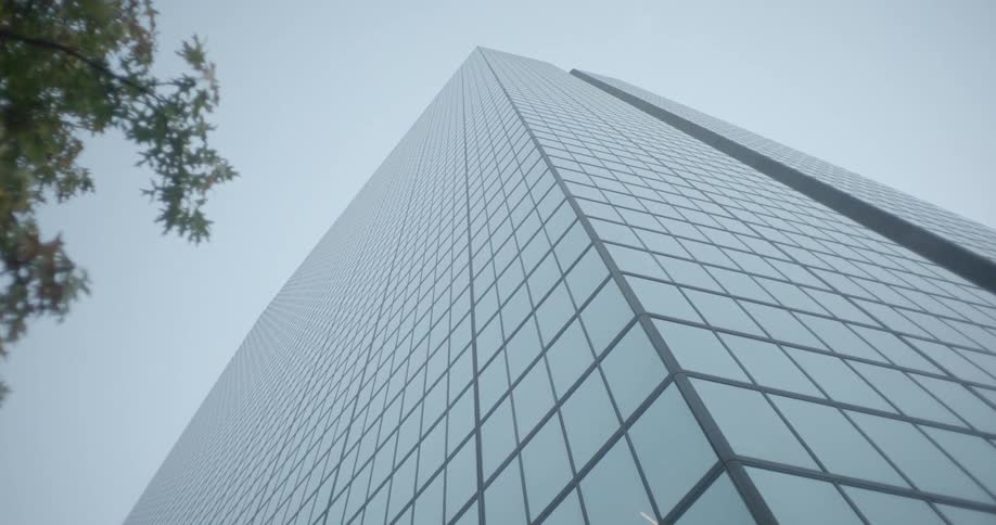 A slow pull-out from a low angle on a modern glass skyscraper reveals its immense height against a plain sky