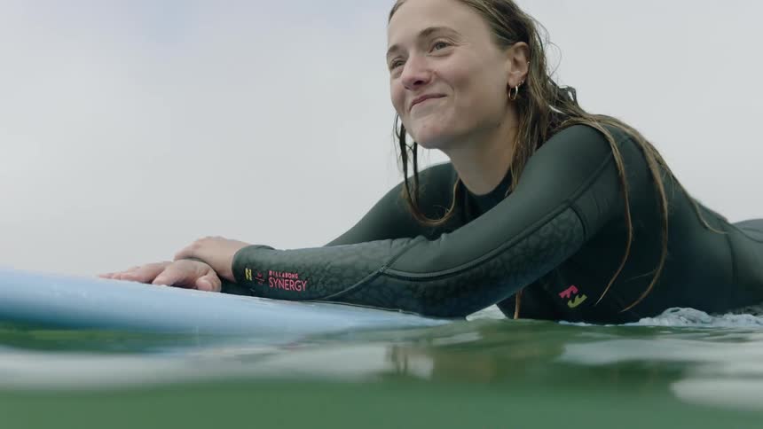 A happy female surfer in a wetsuit smiles while floating on her surfboard in the calm ocean