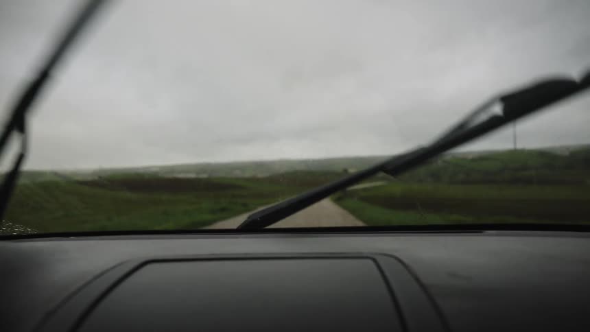 From inside a parked car, windshield wipers clear away the rain, offering a view of a winding rural road on a gloomy day
