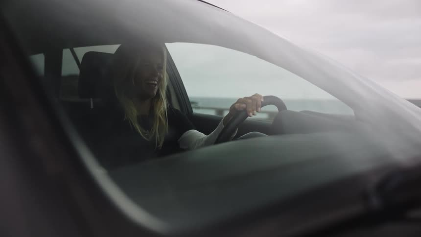 A happy young woman with blonde hair sits in the driver's seat of her car and smiles while parked near the ocean