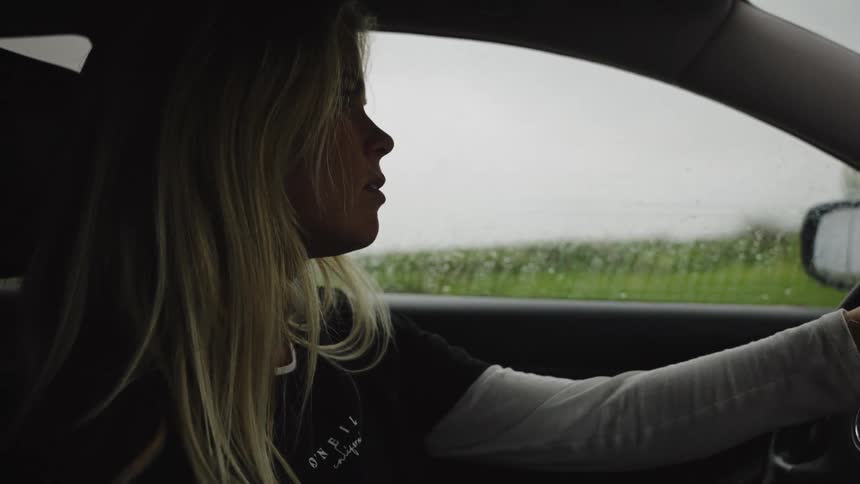 A slow motion side profile shot captures a pensive young woman driving her car on a rainy day through the countryside