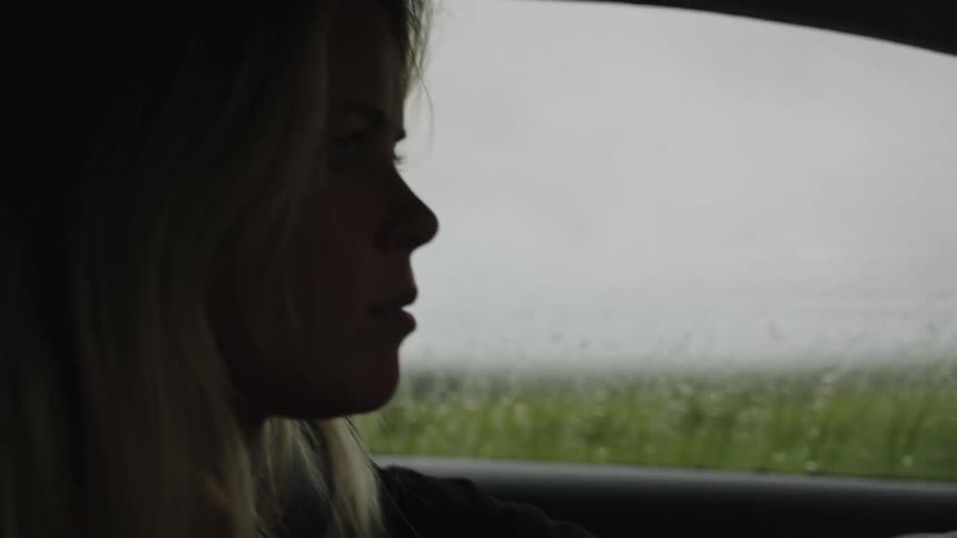 A slow-motion close-up of a silhouetted woman driving her car on a rainy day as the focus shifts from raindrops on the window