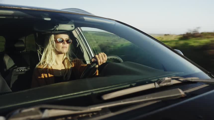 A blonde woman wearing sunglasses drives her car on a scenic road trip through the countryside