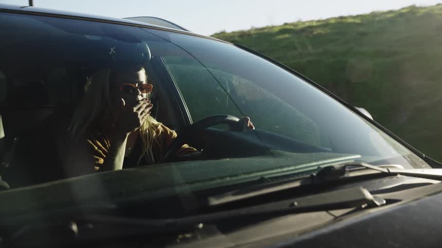 Slow motion shot through a car windshield of a tired woman with sunglasses yawning during a sunny drive in the countryside
