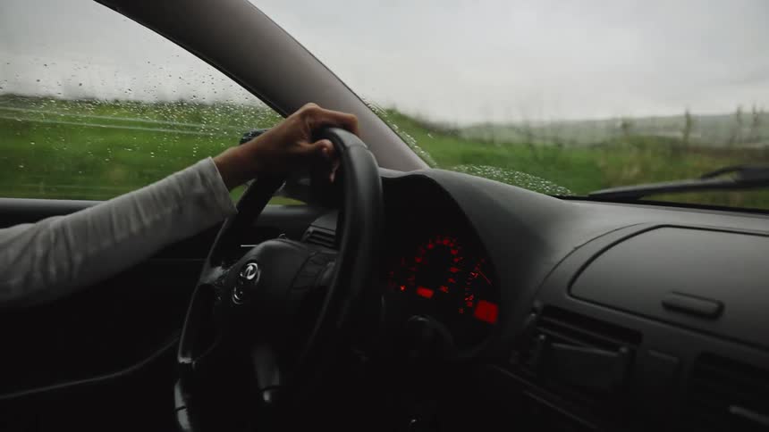 A woman drives a car down a rural road on a rainy day, viewed from the passenger seat