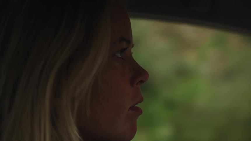 A contemplative blonde woman drives down a rural road, her face illuminated by soft, passing light
