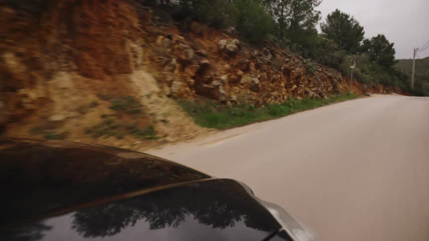 POV shot from the hood of a black car driving along a winding rural road beside a rocky embankment on a cloudy day