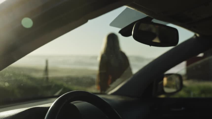 A view from inside a car shows a blurry figure wrapped in a blanket standing in front of the vehicle, looking out at the ocean during golden hour