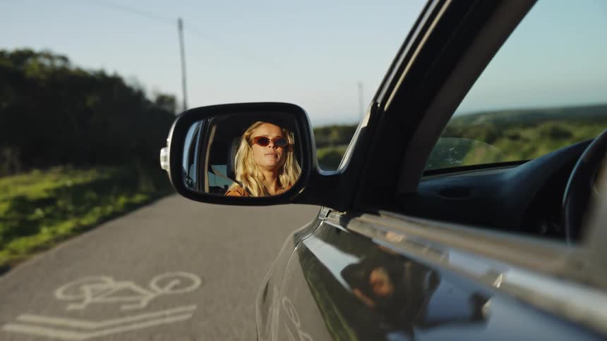 A blonde woman wearing sunglasses is reflected in a car's side-view mirror while driving down a scenic country road at sunset