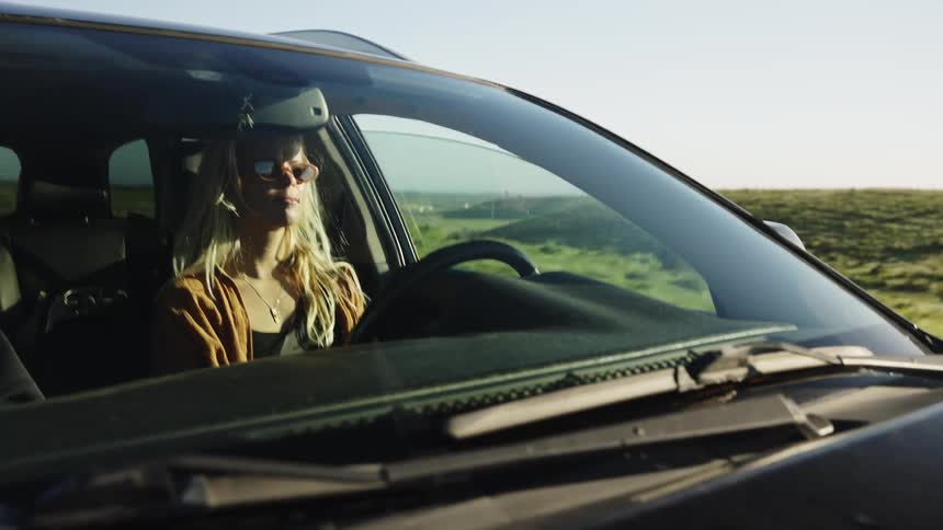 A blonde woman in sunglasses drives her car through the sunny countryside during a road trip