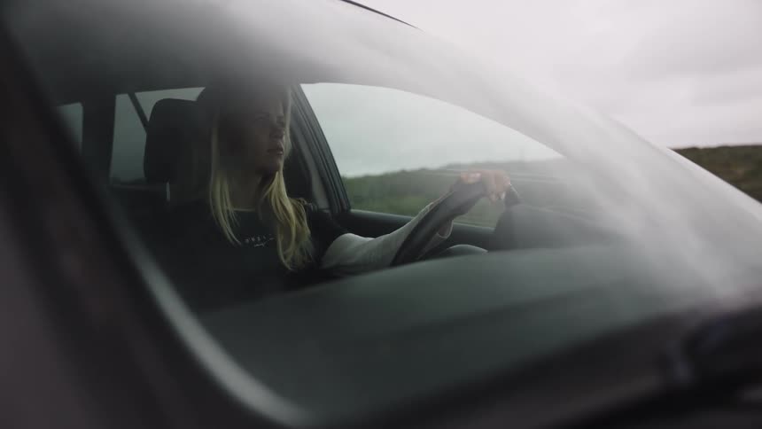 A young woman concentrates on the road ahead as she drives her car through a rural landscape on a cloudy day