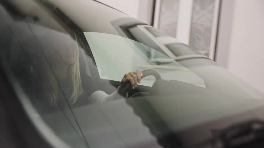 A focused blonde woman maneuvers her car, seen through the front windshield with environmental reflections