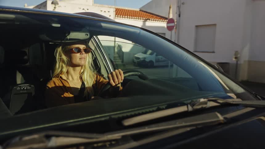 A stylish blonde woman with sunglasses smiles as she drives her car through a sunny town during the golden hour