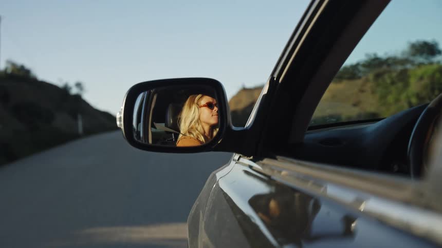 Stylish blonde woman wearing sunglasses is reflected in a car's side-view mirror while driving during a sunset road trip