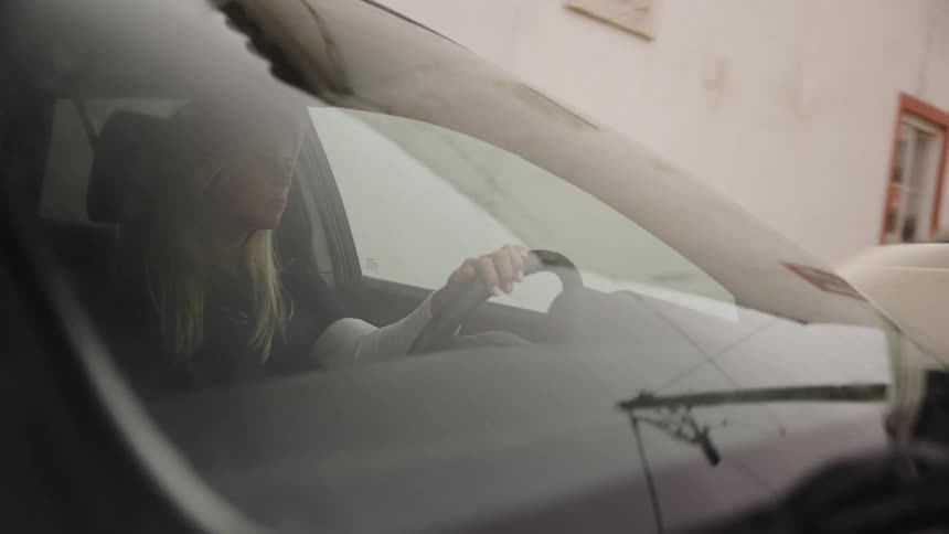 A woman with blonde hair drives a car down a street, shot through the windshield with reflections passing over