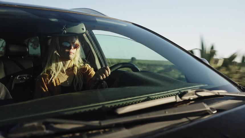 A stylish young woman with blonde hair and sunglasses enjoys a sunny drive through the countryside on a road trip