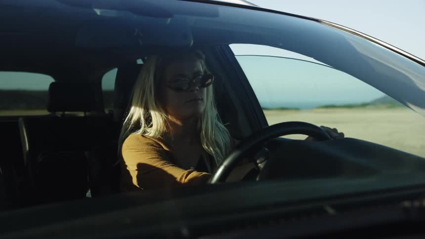 Slow motion shot of a blonde woman in sunglasses driving a car through a sunny desert environment, filmed through the windshield