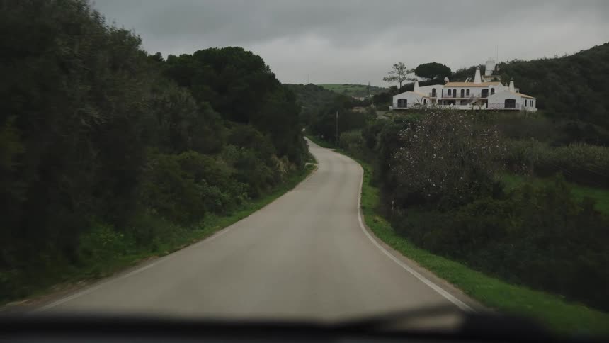 POV shot driving along a narrow, winding country road through green hills towards a distant white house on an overcast day