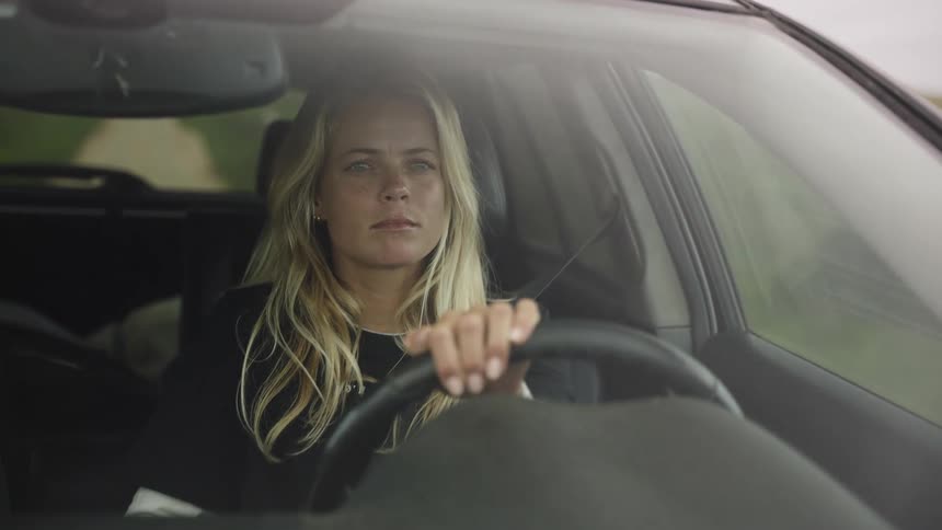 A focused blonde woman with long hair drives her car, as seen from the front windshield