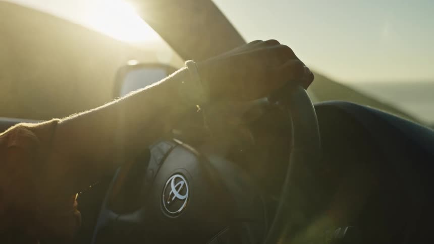 Close-up of a driver's hand on the steering wheel, illuminated by the warm, golden glow of the setting sun on a scenic drive