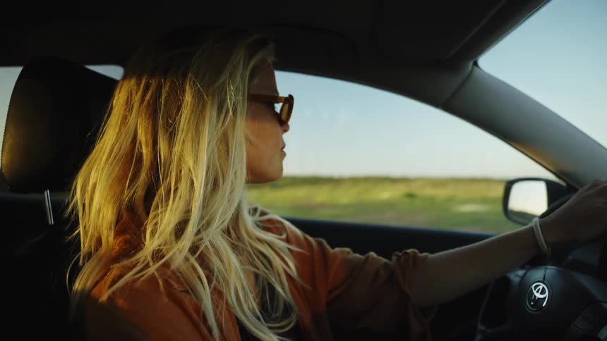 A stylish young blonde woman drives a car through the countryside as the golden hour sun shines on her face
