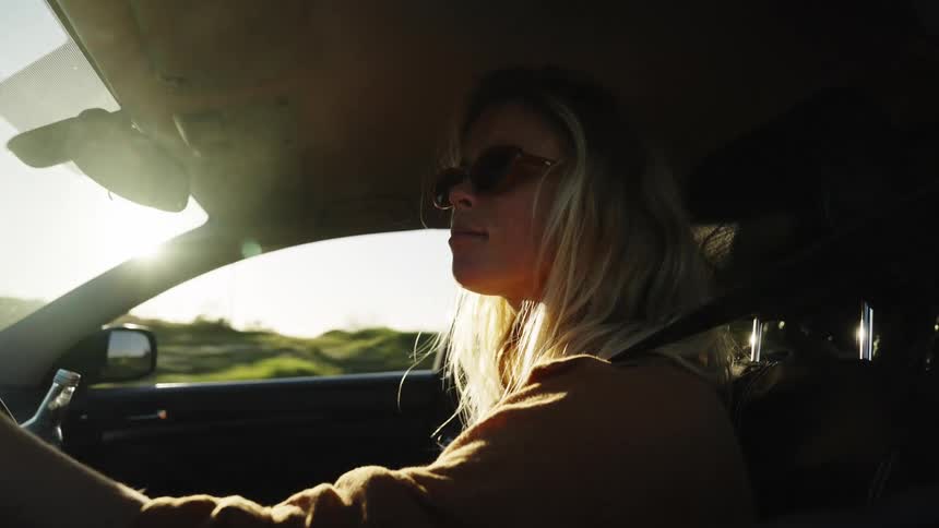 A stylish blonde woman with sunglasses enjoys a drink while driving her car down a scenic road during a beautiful sunset