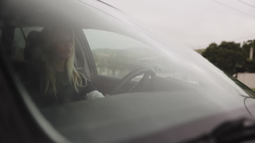 Viewed through the windshield, a young blonde woman navigates her car through town on a cloudy and rainy day