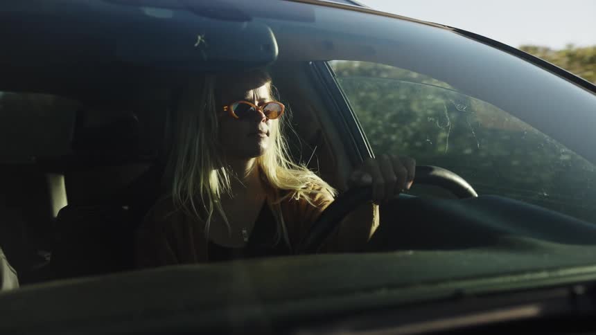 A stylish blonde woman in sunglasses drives a car during the golden hour, seen through the windshield in slow motion