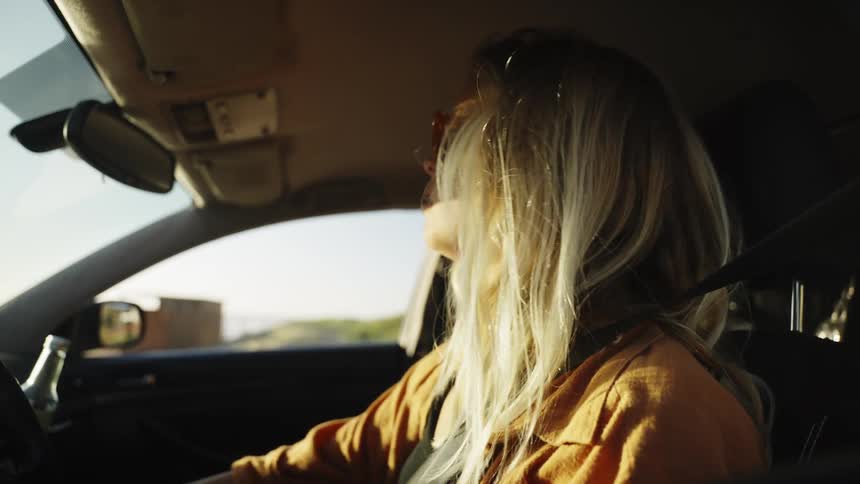 A stylish young blonde woman in sunglasses fastens her seatbelt in her car, ready for a drive at golden hour
