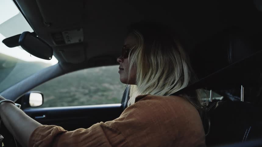 A young blonde woman in sunglasses drives her car along a scenic road during the golden hour in slow motion