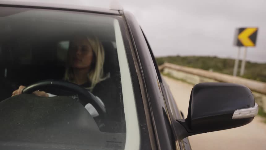 A blonde woman drives a black SUV along a rural road on an overcast day, seen from a front passenger perspective