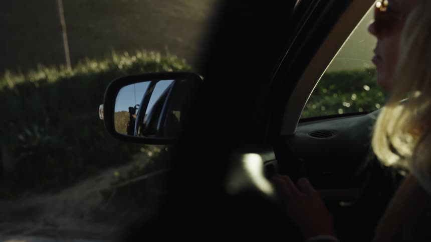 A cool blonde woman's reflection is seen in a car's side-view mirror as she exits the vehicle in the warm golden hour sunlight