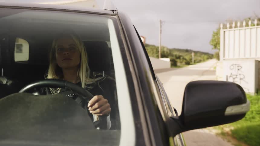 A focused blonde woman drives an SUV down a quiet rural road on an overcast day, filmed from an exterior side view