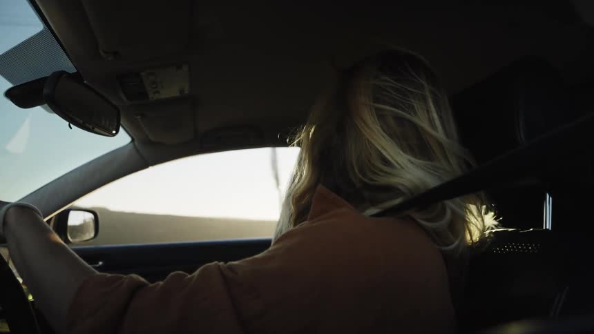 A young woman with sunglasses drives her car down a scenic road during the golden hour, her blonde hair blowing in the wind