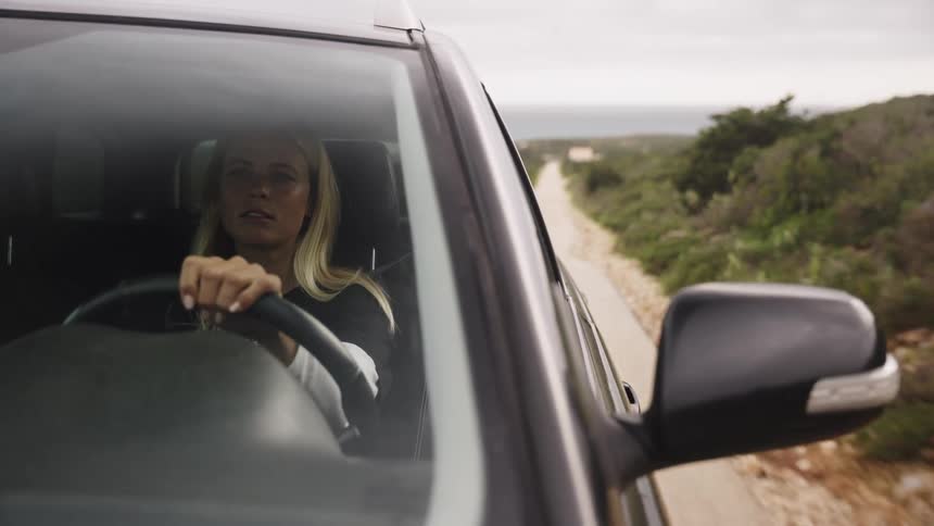 A blonde woman drives her car down a scenic coastal road in slow motion, with the ocean visible in the background