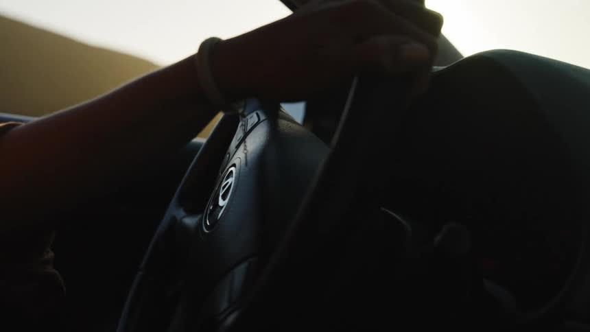 A cinematic close-up shows a driver's hands on the steering wheel, bathed in the warm glow of sunset lens flare