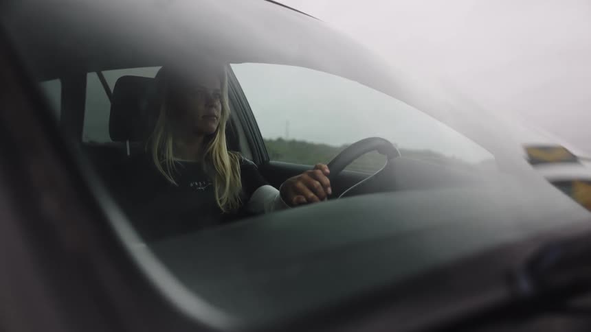 A focused young woman drives down a rural road on an overcast day as seen through the car's windshield