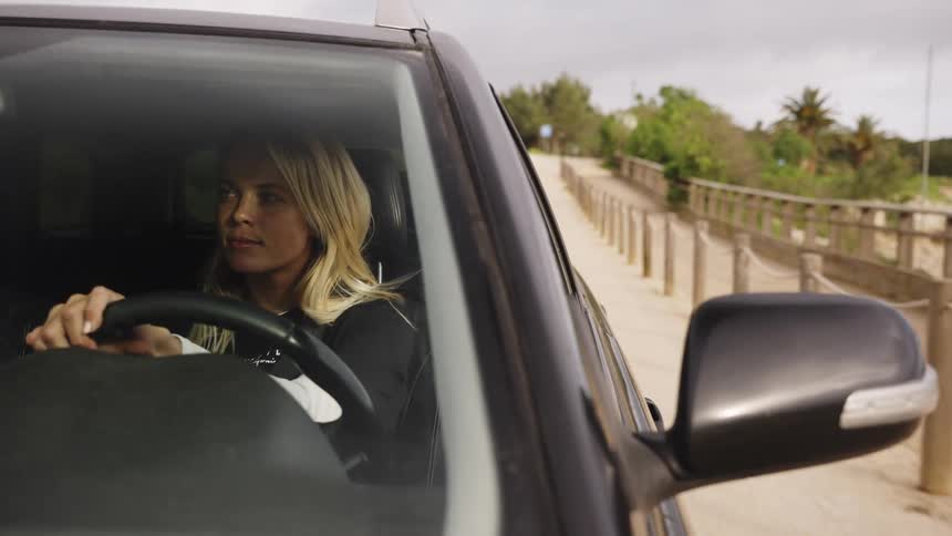 A young blonde woman starts to drive her SUV along a scenic road that runs parallel to a beautiful coastline