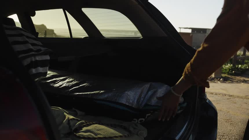 A person opens the trunk of their car and pulls out a surfboard in slow motion, getting ready to surf at the beach