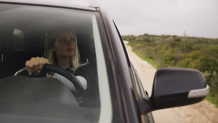 A blonde woman takes a slow, thoughtful drive in her car down a dirt road through the quiet countryside