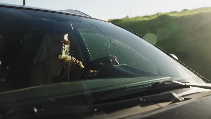 A cool blonde woman in sunglasses drives a car along a scenic country road during a sunny day
