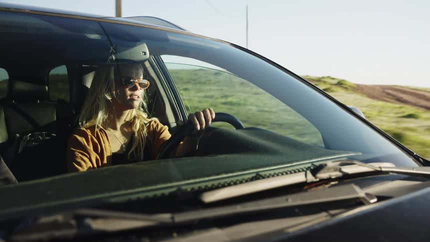 A young blonde woman wearing sunglasses smiles as she drives her car along a scenic country road at sunset