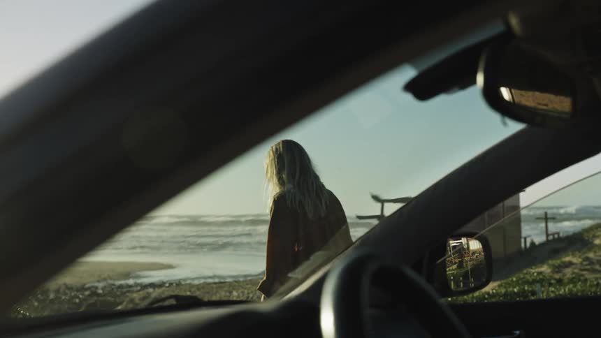 Filmed from inside a car, a woman with blonde hair gets out and stands on the shore, watching the ocean