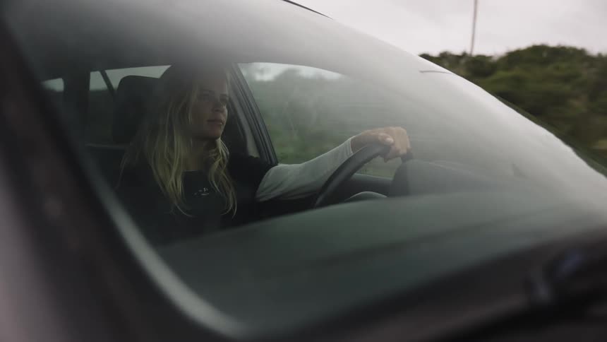 A blonde woman drives her car through the countryside on an overcast day, as seen through the front windshield
