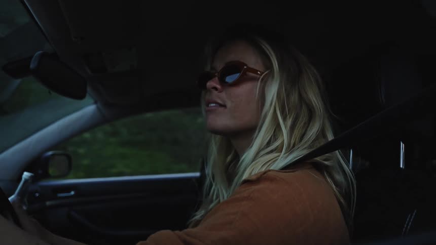 A young blonde woman wearing sunglasses drives her car down a scenic road in slow motion