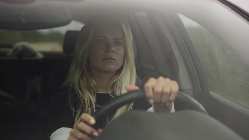 A contemplative young woman with long blonde hair drives her car down a rural road on an overcast day, viewed through the windshield